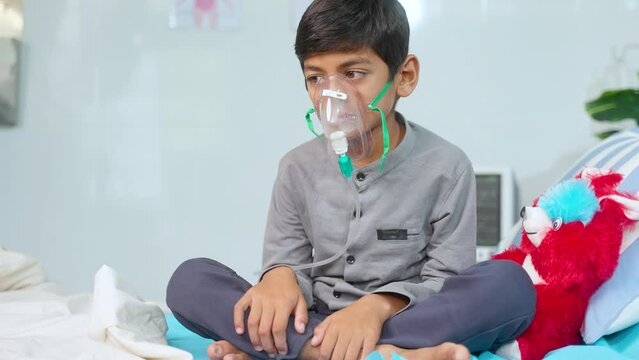 Worried sick kid with oxygen mask sitting on bed at hospital ward - concept of breathing problems, respiratory infection and health care treatment.