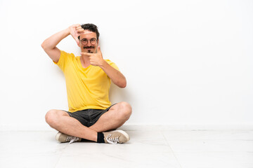 Fototapeta premium Young caucasian man sitting on the floor isolated on white background focusing face. Framing symbol