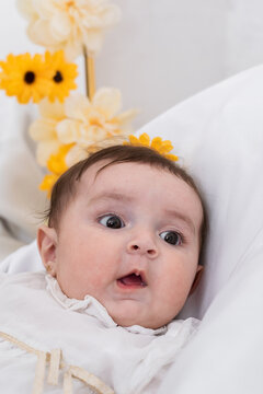 Close-up Of A Latin Baby Girl Lying On Her Crib Looking To The Side With An Amazed Face. Little Girl On A White Background With Yellow And White Flowers.