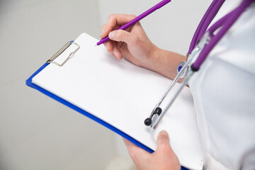 Folder, clipboard with white blank paper sheet in female doctor hands. Purple pen in hand and stethoscope on chest. Copy space