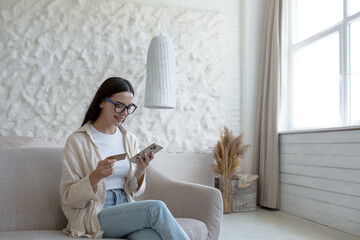Beautiful young woman at home in glasses sitting on the sofa using a mobile phone smartphone for online shopping in a store, holding a bank credit card in her hands
