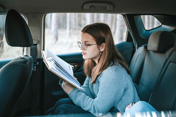 Concentrated caucasian teen girl with loose hair wearing spectacles and knitted grey sweater, sitting inside the car, holding and reading book 