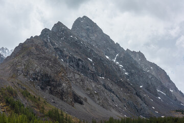 Fototapeta na wymiar Atmospheric mountain landscape with large pointy pinnacle under gray cloudy sky. Gloomy scenery with high pointed mountain peak with sharp rocks in low clouds. Huge rocky peaked top in rainy weather.