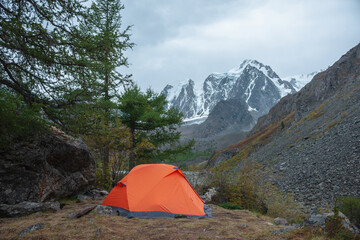 Dramatic landscape with alone orange tent on forest hill among rocks and autumn flora with view to large snowy mountain range under cloudy sky. Lonely tent and fading autumn colors in high mountains.