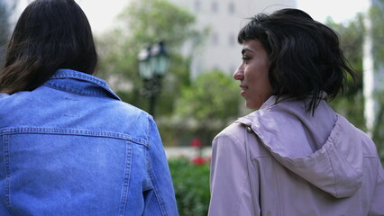 Female friends gossiping outside while walking together. Back of two people talking outdoors