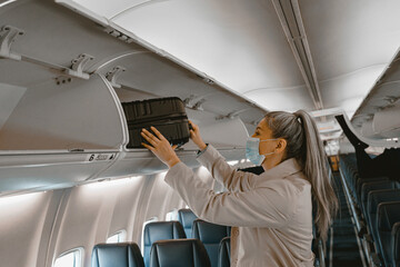 Woman traveler in mask putting luggage into overhead locker on airplane during boarding