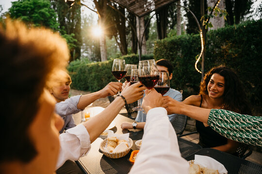 Group of friends outside eat and have fun together toasting with red wine glasses at sunset at the hotel restaurant where are staying - Cheerful people laugh and joke evening  - Party at home