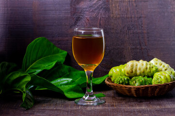 Noni juice in wineglass and noni fruit on basket with green leaf on rustic wooden background. 