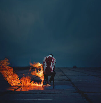 a man covered in blood sits on a burning chair. a man in a bloody white shirt sits on a burning chair. Zombie man. The man is covered in blood and looks like a zombie. Surreal themed photo