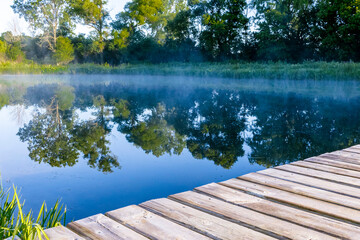 A wooden deck with a steaming pond in the background reflecting wetland plants and trees with a blue sky.