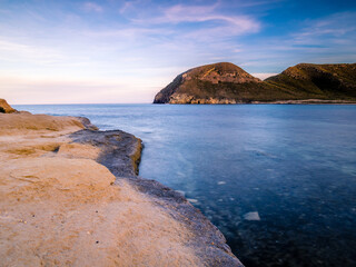 Fotografía de larga exposición en la costa mediterránea con rocas en primer plano y montañas deserticas de fondo de imagen.