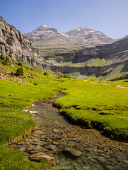 Paisaje alpino donde un riachuelo recorre un valle verde dirección a dos montañas bajo un cielo azul.