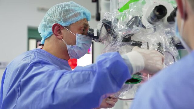 Confident surgeon looks attentively into the binoculars of powerful microscope. Doctor adjusts equipment and takes metal forceps.