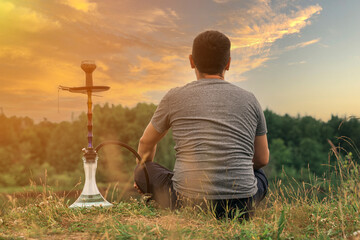 A man sits by the riverbank at sunset and smokes a hookah