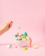 Creative concept of .a bowl full of sweet and tasty candies and gummies from which it is taken with chopsticks as if it were Chinese food. Pastel pink background..