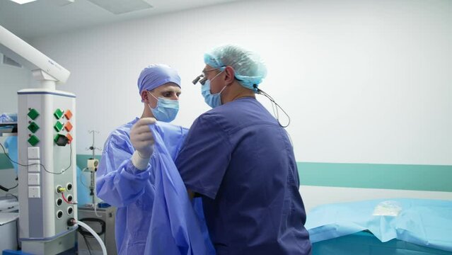 Young male assistant holds the blue robe for the surgeon. Female nurse ties the robe at the doctor's back. Surgeon getting ready for operation.