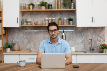 Portrait of handsome man sitting at the table and working on laptop in the kitchen at home