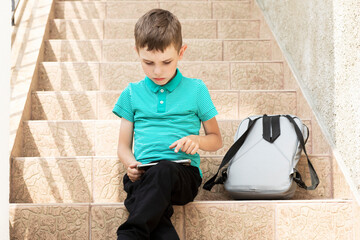 European (American) boy (pupil, student) sits on stairs after elementary school lessons (primary class)and looks at cell phone, playing a game, backpack(school bag) is beside, daytime.Horizontal plane
