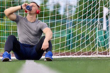 Football goalkeeper sits on green surface in center of goal, drinking water from sports bottle after game. Young goalkeeper rests after training, quenches thirst, listens music in modern headphones © Aleksandr