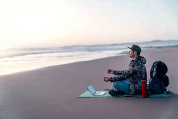 mature digital nomad sitting on the beach meditating with his laptop on the shore of the beach - obrazy, fototapety, plakaty