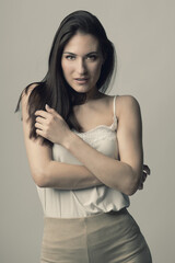 Fototapeta premium Fashion, style, beauty and make-up concept. Beautiful woman fashion studio portrait with long dark hair and day make-up with seductive look and pose with crossed arms. Toned image with beige color