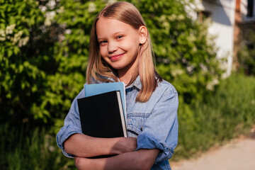 Fototapeta premium Smiling girl holding notebooks and looking at camera outdoor. Child ready for school. Back to school concept