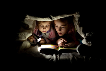 two sisters are reading a book under the covers at night with a flashlight - obrazy, fototapety, plakaty