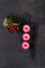 Cherry mousse dessert with curly jelly, in the form of round sweet sushi, sprinkled with colored coconut shavings, on a serving board. Dark gray background. Top view
