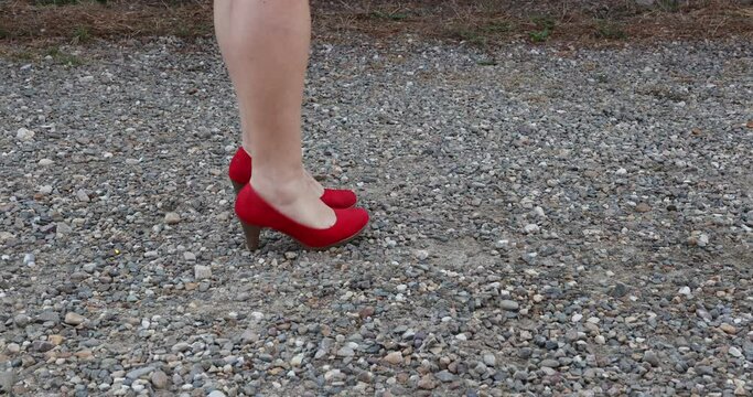 Female adult legs wearing elegant red high-heeled shoes on the feet, walking on a stony path, stopping to adjust the shoe, and awkwardly walking again. Concept of elegance in difficult times