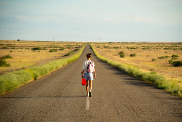 Fototapeta premium A girl walks along an empty road in the steppe. She has a ukulele in her hands and a backpack on her shoulders. It's summer and warm outside. Open road.