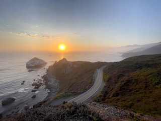 Golden sunset over State Route 1, on the Pacific Ocean. Pacific Coast Highway, Central California near Gorda. Slightly foggy, golden sunset with a few clouds over the pacific ocean. Curving Highway