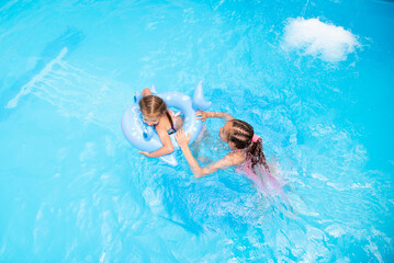 Two sister girls of 11-13 and 6 years old swim in a pool with blue water and have a fan. The older girl has african braids braided with zi-zi ribbons. Summer. Family vacation.