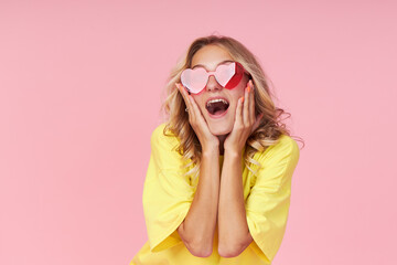 Portrait of an excited young woman in heart-shaped glasses, yellow t-shirt on a pink background. Surprised girl looks to the left with her eyes wide open