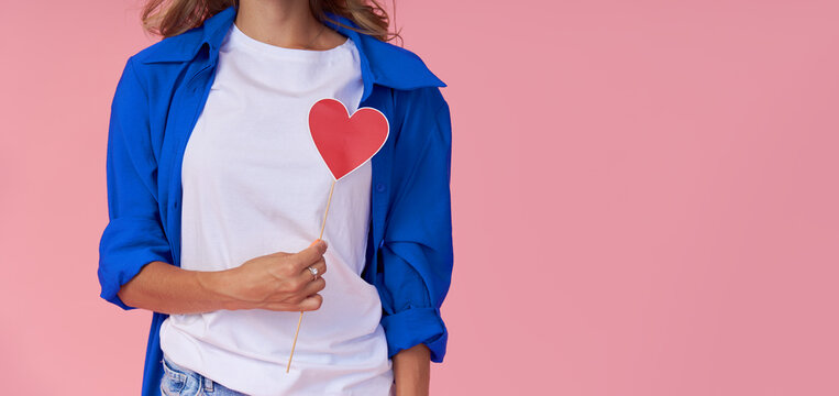 A young woman in a blue shirt holds a red paper heart in front of her chest. Pink studio background. The concept of women's health and diseases of the cardiovascular system
