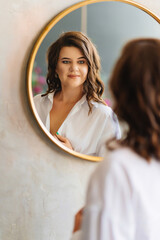 a brunette woman in a white shirt looks at her reflection in a round mirror.