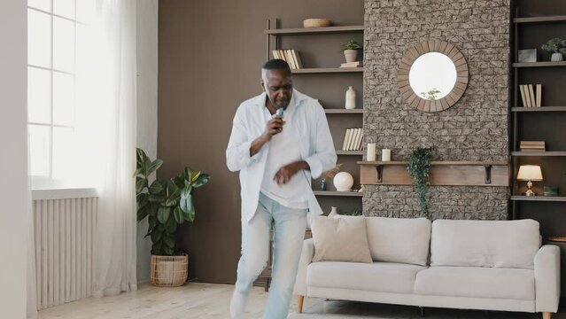 Mature crazy artistic african american man energetic vocalist singer grandpa homeowner using mobile phone as microphone pretending singing in karaoke moving break-dance enjoying domestic party at home
