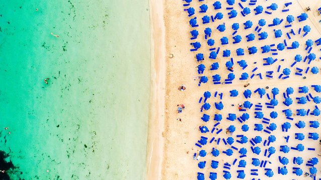 Aerial bird's eye view of Makronissos organised beach coastline, Ayia Napa, Famagusta, Cyprus from above. Blue aligned umbrellas, golden sand, parasols, people sunbathing sun beds clean turquoise sea.