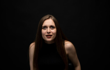 Fototapeta na wymiar Portrait of upset young beautifil brunette girl with long hair in a black t-shirt in a black room. Emotion of angry and upset.