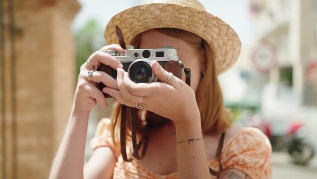 Young redhead woman tourist wearing summer hat using vintage camera at street