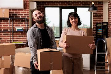 Portrait of married happy couple carrying boxes to move in apartment property, using furniture storage to decorate family home. Enjoying household relocation to start new beginnings.