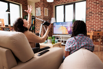 Angered woman screaming at sky because of lost game round of videogame played on gaming console. Diverse people sitting on sofa while playing space simulator game on modern entertainment device.