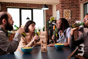 Multiethnic friends looking at each other while sipping beer and playing with wood blocks. Diverse people sitting together at home in living room while talking and enjoying fun leisure activity.
