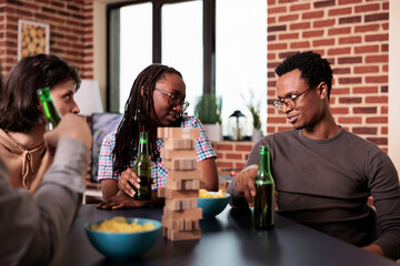 Diverse group of close friends sitting at table in living room while having fun together with society games. Casual people sitting at home while enjoying fun leisure time activities with wooden blocks