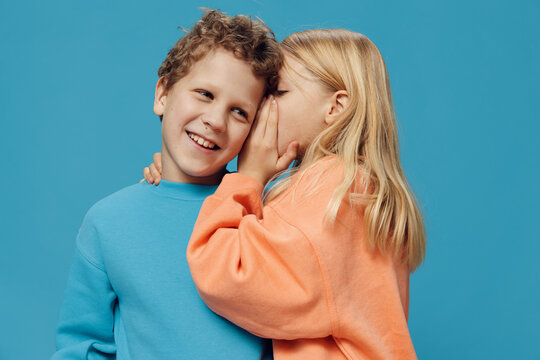  Children Of School Age In Colored Sweaters Stand On A Blue Background And The Girl Tells The Boy A Secret In The Ear While Standing Sideways To The Camera. The Theme Of Friendship Between Children