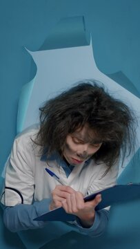 Vertical video: Mad goofy scientist taking notes on papers after dangerous explosion, acting crazy and doing funy foolish facial expressions. Fun amusing chemist being insane and having wacky hair.