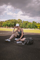Beautiful Caucasian girl athlete dressed in sportswear: shorts and top, sneakers with cap and bag sitting on stadium in summer. Happy female outdoor sport, lifestyle concept. Copy space for text