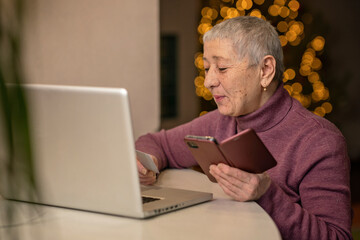 An elderly woman sitting in front of a laptop holds a bank card in her hands and makes online purchases.