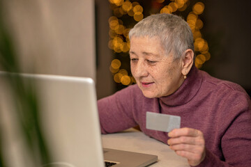 An elderly woman sitting in front of a laptop holds a bank card in her hands and makes online purchases.