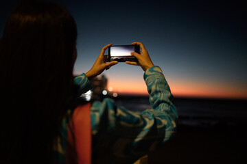 Trendy tourist taking photo on her phone of the ocean with beautiful sunset on the horizon at night outdoors. Influencer or vlogger taking picture of nature sea view to post or share on social media - obrazy, fototapety, plakaty