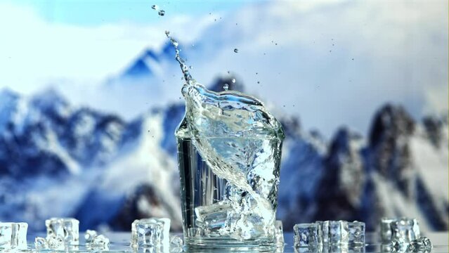 An ice cube falls into a glass of water with splashes. Against the backdrop of mountains. Filmed is slow motion 1000 fps.